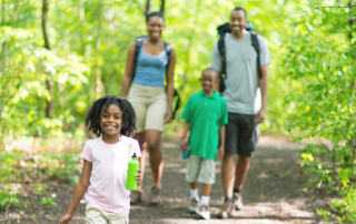 A happy family of four walking on a trail in the woods surrounded by green trees. Mom and dad have backpacks on and the young daughter is carrying a water bottle.