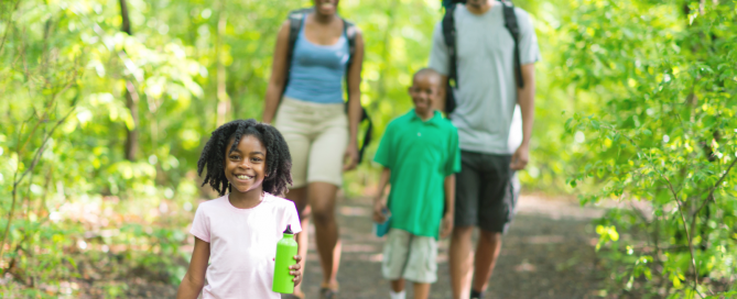 A happy family of four walking on a trail in the woods surrounded by green trees. Mom and dad have backpacks on and the young daughter is carrying a water bottle.