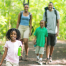 A happy family of four walking on a trail in the woods surrounded by green trees. Mom and dad have backpacks on and the young daughter is carrying a water bottle.