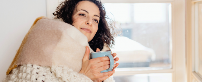 woman wrapped in a cozy blanket and holding a blue mug of a warm drink, smiling softly to depict a cozy setting at home