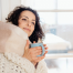 woman wrapped in a cozy blanket and holding a blue mug of a warm drink, smiling softly to depict a cozy setting at home