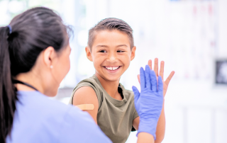 image of young boy at a doctors office and giving a high five to his provider. He is smiling big and we see the back of the provider and her hand in a medical glove.