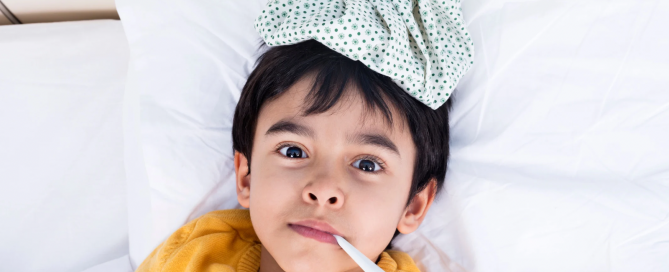 image of a young boy around 5 laying in bed with an ice pack on his head and a thermometer in his mouth to portray a sick child