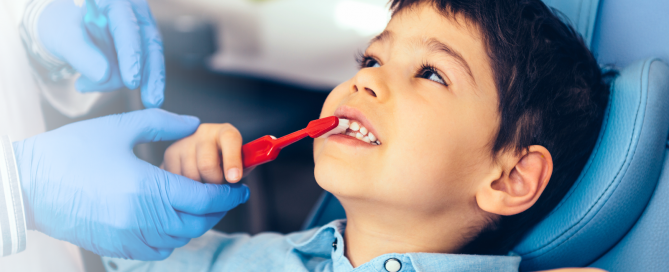 Image of young boy in a dental chair, being assisted in brushing his teeth with a red toothbrush. He is calm and half smiling