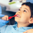 Image of young boy in a dental chair, being assisted in brushing his teeth with a red toothbrush. He is calm and half smiling